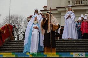 Kobieta i mężczyzna przebrani za Maryję i Józefa stoją na schodach. Mężczyzna przemawia do mikrofonu.