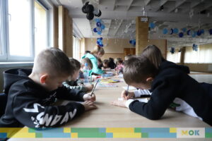 Grupa dzieci siedzący przy stołach w trakcie zajęć.