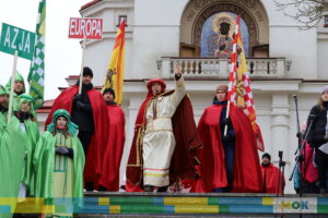 W środku kadru mężczyzna uniesioną ręką w górze pozdrawia innych ludzi.