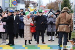 W środku kadru stoi kobieta i mężczyzna, który przechyla kubek.