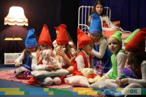 Grupa dzieci przebrane w kolorowe stroje siedzi na scenie.