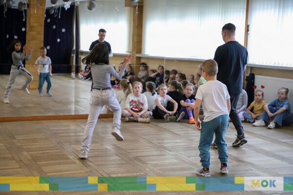 Dziewczynka i chłopiec wykonują układ taneczny pod nadzorem dorosłego instruktora.