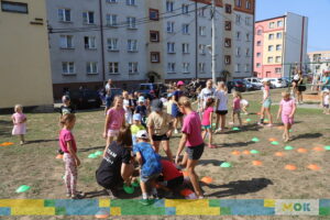 Dzieci uczestniczą w zabawie na placu, ustawione w kręgu i wykonujące różne aktywności ruchowe. W tle widać bloki mieszkalne oraz obserwujących dorosłych.