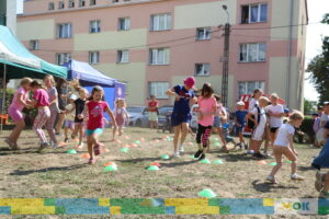 Dzieci biorą udział w dynamicznej zabawie, biegając między ustawionymi na trawie pachołkami. W tle widoczne są namioty organizacyjne oraz bloki mieszkalne. Zabawa wywołuje wiele radości i śmiechu.