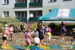 Dzieci biorą udział w zabawie na świeżym powietrzu, otoczone kolorowymi piłeczkami, które rozsypują się z dużego przezroczystego pojemnika trzymanego przez instruktorkę. Dzieci wyglądają na podekscytowane i chętnie uczestniczą w zabawie.