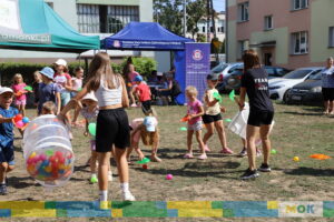 Dzieci zbierają kolorowe piłeczki rozrzucone na trawie, podczas gdy animatorka nadzoruje zabawę. Na tle zielonego namiotu widać inne osoby zaangażowane w wydarzenie.