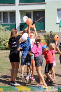 Grupa dzieci wyciąga ręce, próbując złapać pomarańczowy stożek, który trzyma instruktorka. Dzieci z zaangażowaniem uczestniczą w grze, śmiejąc się i rywalizując o zdobycie stożka.