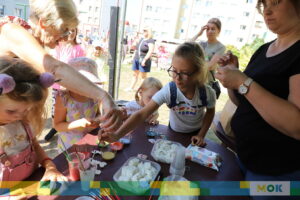 Kilkoro dzieci i dorosłych bawi się przy stole z materiałami plastycznymi. Dzieci kolorują figurki z masy solnej, używając pędzli i różnych farb. Opiekunowie pomagają i nadzorują ich prace.