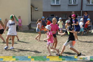 Dzieci biegają po placu, trzymając różnokolorowe szarfy lub kawałki materiałów, angażując się w energiczną zabawę ruchową na świeżym powietrzu. W tle widać dorosłych oraz budynki mieszkalne.