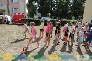 Grupka dzieci ustawionych w szeregu na trawniku, biorąca udział w aktywności ruchowej. Niektóre z dzieci mają na sobie specjalne okulary, co może sugerować zabawę z elementem utrudniającym widzenie. W tle widać dorosłych obserwujących zabawę oraz zaparkowane pojazdy.