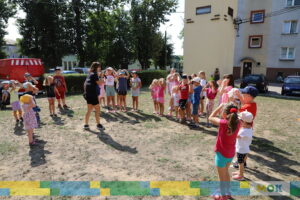 Dzieci stoją w kole na otwartej przestrzeni, wykonując ćwiczenia ruchowe pod okiem instruktorki, która trzyma mikrofon. Dzieci mają ręce na głowie, a w tle widać bloki mieszkalne oraz zaparkowane samochody.