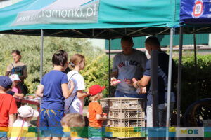 Mężczyźni pod zielonym namiotem rozdają dzieciom i dorosłym różne smakołyki lub nagrody. Dzieci w kolorowych czapkach z radością odbierają drobne upominki.