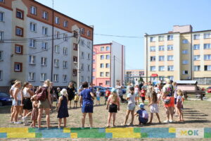 Grupa uczestników zgromadzona na placu przed budynkami mieszkalnymi. Dorośli i dzieci stoją w kręgu, przygotowując się do zabawy lub aktywności ruchowej.