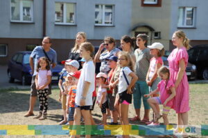 Dzieci i dorośli stoją razem, uważnie słuchając prowadzącego wydarzenie na świeżym powietrzu. Wszyscy są skupieni na tym, co dzieje się przed nimi, a w tle widać bloki mieszkalne.