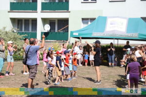 Dzieci i dorośli stoją w kole przed instruktorką, która prowadzi zajęcia. Grupa entuzjastycznie wykonuje ruchy rękami. Na namiocie w tle widnieje logo „kulturamonki.pl”.