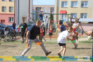 Dorosły mężczyzna i dziecko biorą udział w zabawie na trawie, gdzie biegają, trzymając pomarańczowe pompony. W tle widoczny jest plac zabaw oraz dzieci zaangażowane w różne aktywności.