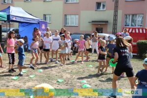 Grupa dzieci bierze udział w zajęciach plenerowych, wykonując różne zadania ruchowe na świeżym powietrzu. Na pierwszym planie widoczna jest instruktorka w czarnej koszulce z napisem „TEAM”, która trzyma zielony dysk. Dzieci śmieją się i angażują w aktywności.