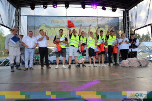 Uczestnicy festiwalu na scenie, w tym laureaci w odblaskowych kamizelkach, podnoszą ręce w geście zwycięstwa. Organizatorzy stoją obok nich, a cała scena oświetlona jest kolorowymi reflektorami.