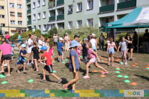 Grupa dzieci i dorosłych bierze udział w różnych aktywnościach na otwartym terenie między blokami. Kolorowe namioty z logo organizatorów i dzieci w kapeluszach tworzą żywą, radosną atmosferę wydarzenia.