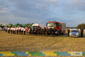 Strażacy, ratownicy medyczni i policjanci stoją w rzędzie na tle wozów strażackich i policyjnych podczas ceremonii na festynie. Każdy z nich jest w służbowym uniformie, gotowy do pokazowego wystąpienia.