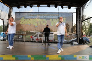 Dwóch wokalistów na scenie śpiewających podczas występu, jeden z mikrofonem w ręku. W tle znajduje się baner „Święto Wsi”, a muzycy w tle grają na instrumentach.