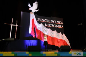 łoda dziewczynka śpiewa na scenie, trzymając mikrofon. W tle duża biało-czerwona flaga oraz napis „JESZCZE POLSKA NIE ZGINĘŁA” z wizerunkiem białego orła na górze i drewnianym krzyżem po lewej stronie.