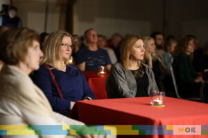 Widok na publiczność podczas wydarzenia. Kilka osób siedzi przy stołach z czerwonymi obrusami, skupionych na obserwacji występu. Na stołach zapalone świeczki.