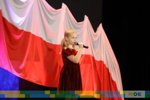 Młoda dziewczynka w czerwonej sukience śpiewa na scenie, trzymając mikrofon. W tle biało-czerwona flaga nadająca scenie patriotyczny charakter.