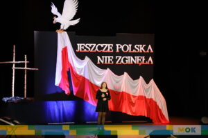 Dziewczyna w czarnej sukience i z biało-czerwoną rozetką śpiewa na scenie przed patriotycznym tłem składającym się z flagi Polski i napisu „JESZCZE POLSKA NIE ZGINĘŁA”.