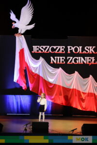 Dziewczynka śpiewa do mikrofonu na scenie, na tle drapowanej flagi Polski i napisu „Jeszcze Polska nie zginęła,” co nadaje występowi patriotycznego charakteru.