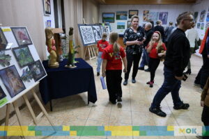 Sala wystawowa z pracami plastycznymi na sztalugach. Dzieci i dorośli spacerują, oglądając eksponaty, w tym rzeźby i obrazy.