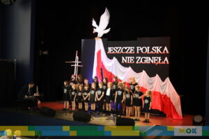 Grupa dzieci ubranych w czarne stroje z motywem orła, stojących na scenie przed dużą biało-czerwoną flagą i napisem "JESZCZE POLSKA NIE ZGINĘŁA". Po lewej stronie pianista grający na klawiaturze.