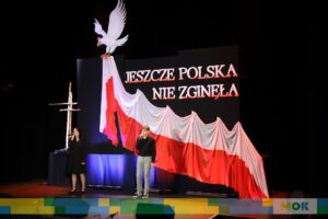 Dwie osoby wykonujące pieśni patriotyczne na scenie podczas przeglądu, jedna z nich śpiewa, a druga trzyma mikrofon. W tle zawieszona jest duża flaga Polski, obok niej krzyż z drewna, a nad flagą widnieje biały orzeł oraz napis "Jeszcze Polska nie zginęła".