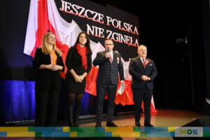 Grupa czterech dorosłych osób na scenie, przemawiających do publiczności w trakcie przeglądu pieśni patriotycznych. Na tle znajduje się duża flaga Polski oraz hasło „Jeszcze Polska nie zginęła” – prowadzący i organizatorzy wydarzenia.