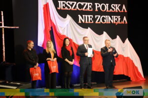 Grupowe zdjęcie uczestników na scenie podczas uroczystości patriotycznej. W tle widać rozpostartą biało-czerwoną flagę oraz napis „Jeszcze Polska nie zginęła”. Osoby na scenie trzymają dyplomy oraz torby z symbolami lokalnymi.