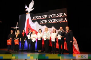 Grupowe zdjęcie na scenie podczas uroczystości patriotycznej. W tle widać rozpostartą biało-czerwoną flagę oraz napis „Jeszcze Polska nie zginęła”.