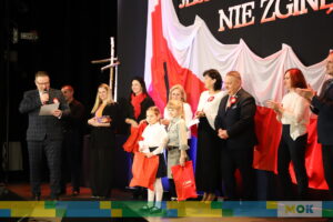 Wręczenie nagród młodym uczestnikom wydarzenia patriotycznego. Mężczyzna w garniturze pochyla się, aby przekazać nagrody dzieciom trzymającym czerwone torby z napisem „I ❤️ Mońki”. W tle znajduje się biało-czerwona flaga.