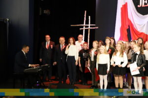 Grupa dzieci i dorosłych śpiewa na scenie podczas patriotycznej uroczystości. Jeden z mężczyzn gra na keyboardzie, a na ścianie wisi duża biało-czerwona flaga z napisem „Jeszcze Polska nie zginęła” i figurą orła.