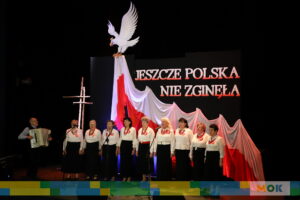 Grupa starszych kobiet ubranych w białe bluzki i ciemne spódnice, śpiewających na scenie przed patriotycznym tłem z biało-czerwoną flagą. Po lewej stronie mężczyzna gra na akordeonie.