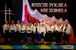 Chór mieszany składający się ze starszych kobiet i mężczyzn w białych koszulach ozdobionych czerwonymi rozetkami. Śpiewają wspólnie pieśni patriotyczne na scenie ozdobionej flagą i napisem "JESZCZE POLSKA NIE ZGINĘŁA".