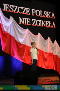 Chłopiec w białej koszulce trzyma mikrofon i śpiewa na scenie przed dużą biało-czerwoną flagą i patriotycznym napisem "JESZCZE POLSKA NIE ZGINĘŁA".