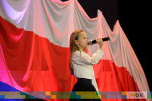 Dziewczynka w białej bluzce i czarnej spódnicy śpiewa do mikrofonu na tle biało-czerwonej flagi, przygotowanej na patriotyczne wydarzenie.